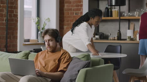 Niño-Trabajando-En-Una-Computadora-Portátil-Sentado-En-Un-Sofá-Mientras-Sus-Compañeros-De-Cuarto-Limpian-La-Cocina-1