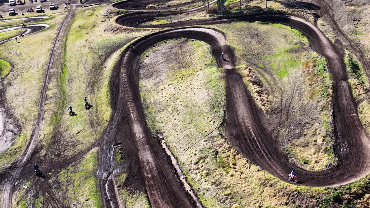 Multiple riders navigate winding dirt motocross track in daylight, aerial perspective, natural outdoor setting