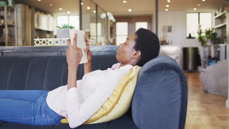 Feliz-Mujer-Afroamericana-Tumbada-En-El-Sofá-De-La-Sala-De-Estar,-Leyendo-Un-Libro