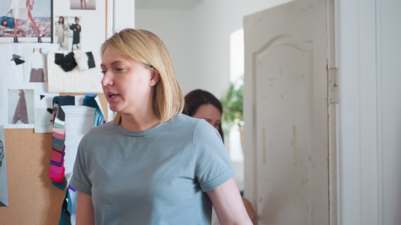 Fashion designer enters workshop with student, picks up paper to explain clothing design ideas surrounded by dummies, fabric samples, and inspiring creative materials in busy fashion studio