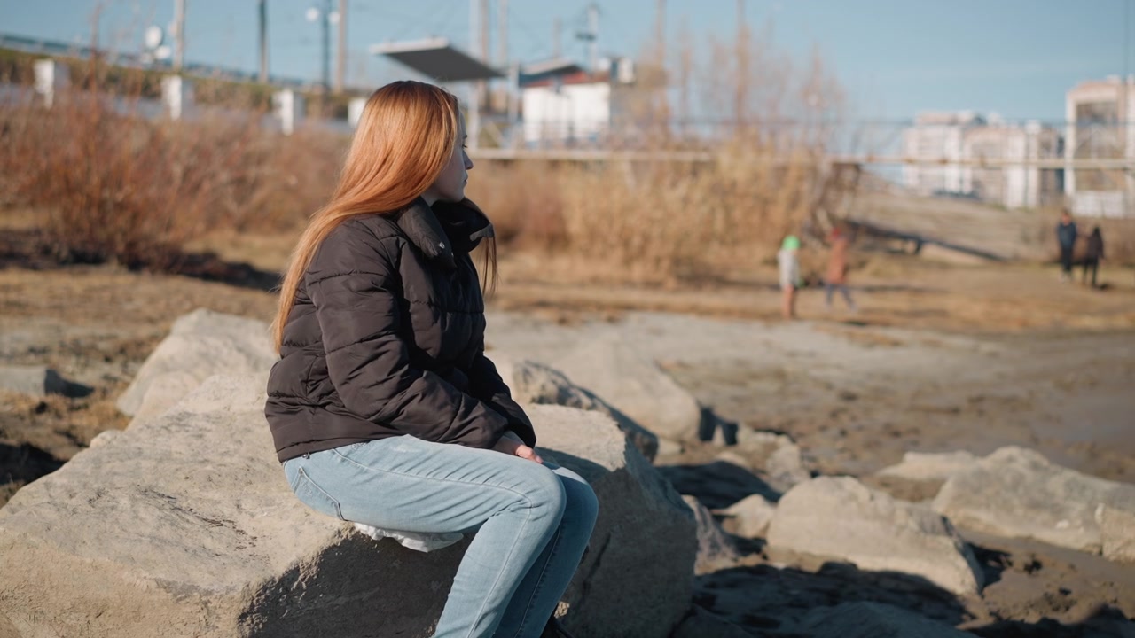 Woman with long hair sits on large rock in outdoor setting wearing black puffer jacket and light blue jeans. background includes dry grassy areas distant buildings construction structures