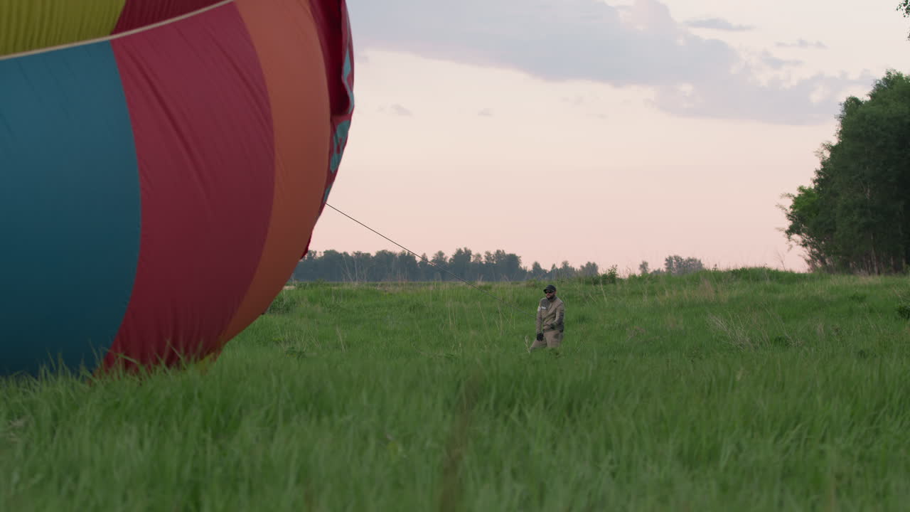 Close up colorful hot air balloon envelope panels glowing under pastel sky as crew member pulls tether rope, vibrant fabric resting on green grassy field with distant tree line backdrop