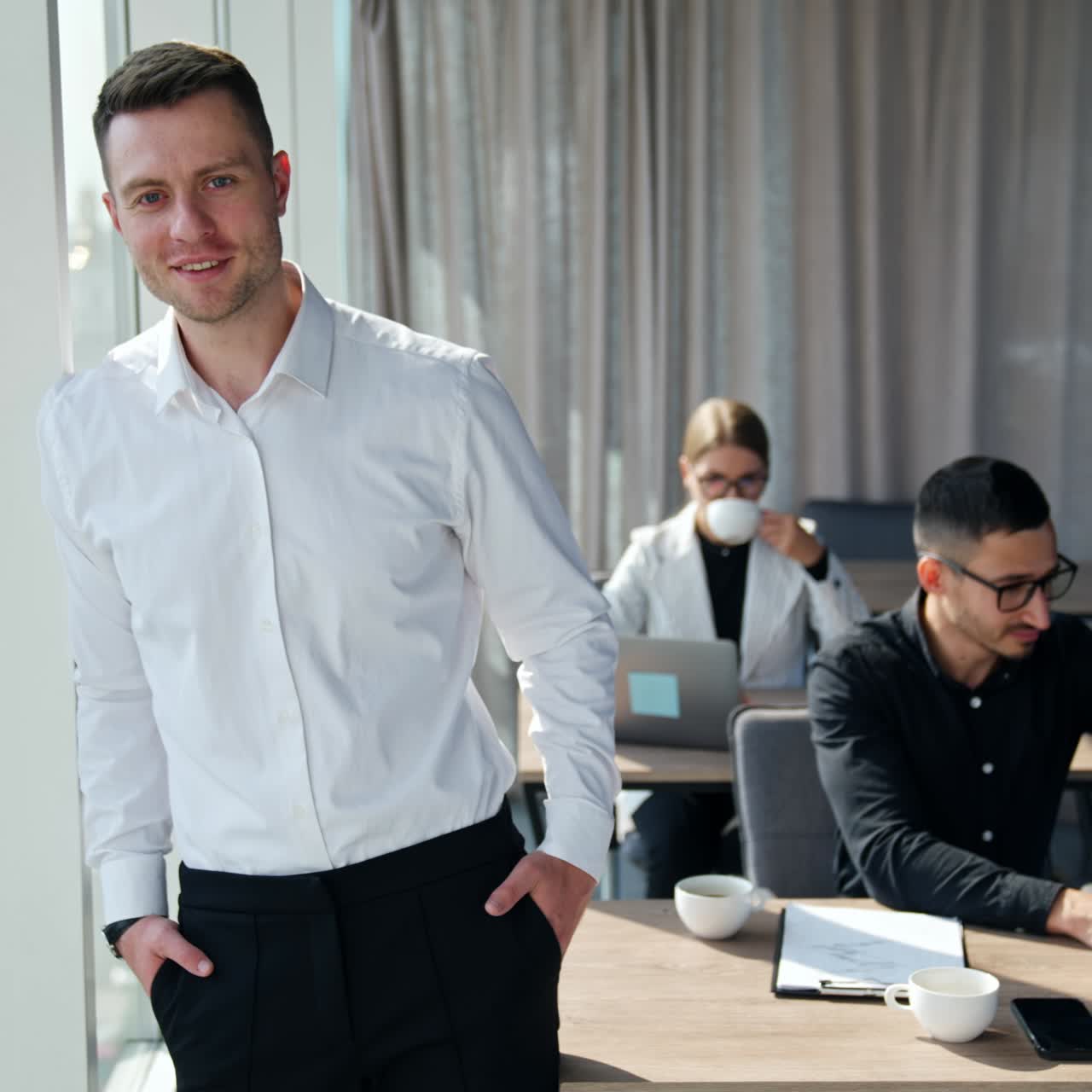 Young handsome businessman wearing white shirt and trousers stands at window smiling. Male and female colleagues working at laptops at their desks