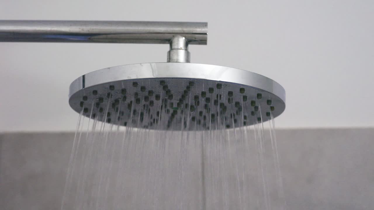 A modern chrome shower head releasing a steady stream of water in a clean, minimal bathroom environment. The close-up view highlights the sleek design and gentle water flow