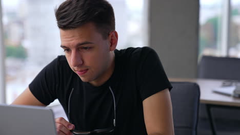 dark-haired handsome man takes off his glasses to look closer at computer screen. freelancer working on laptop in big light room. blurred backdrop.