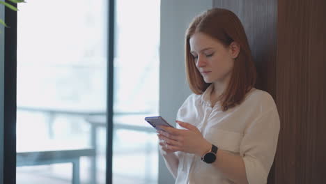 Mujer-Joven-Con-Teléfono-Inteligente-En-La-Oficina-Durante-La-Jornada-Laboral-Enviando-Mensajes-Y-Viendo-Las-Redes-Sociales