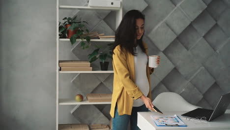 Young happy pregnant business woman uses phone, takes a break stroking her belly while having a hot drink