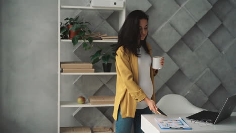 young happy pregnant business woman uses phone, takes a break stroking her belly while having a hot drink