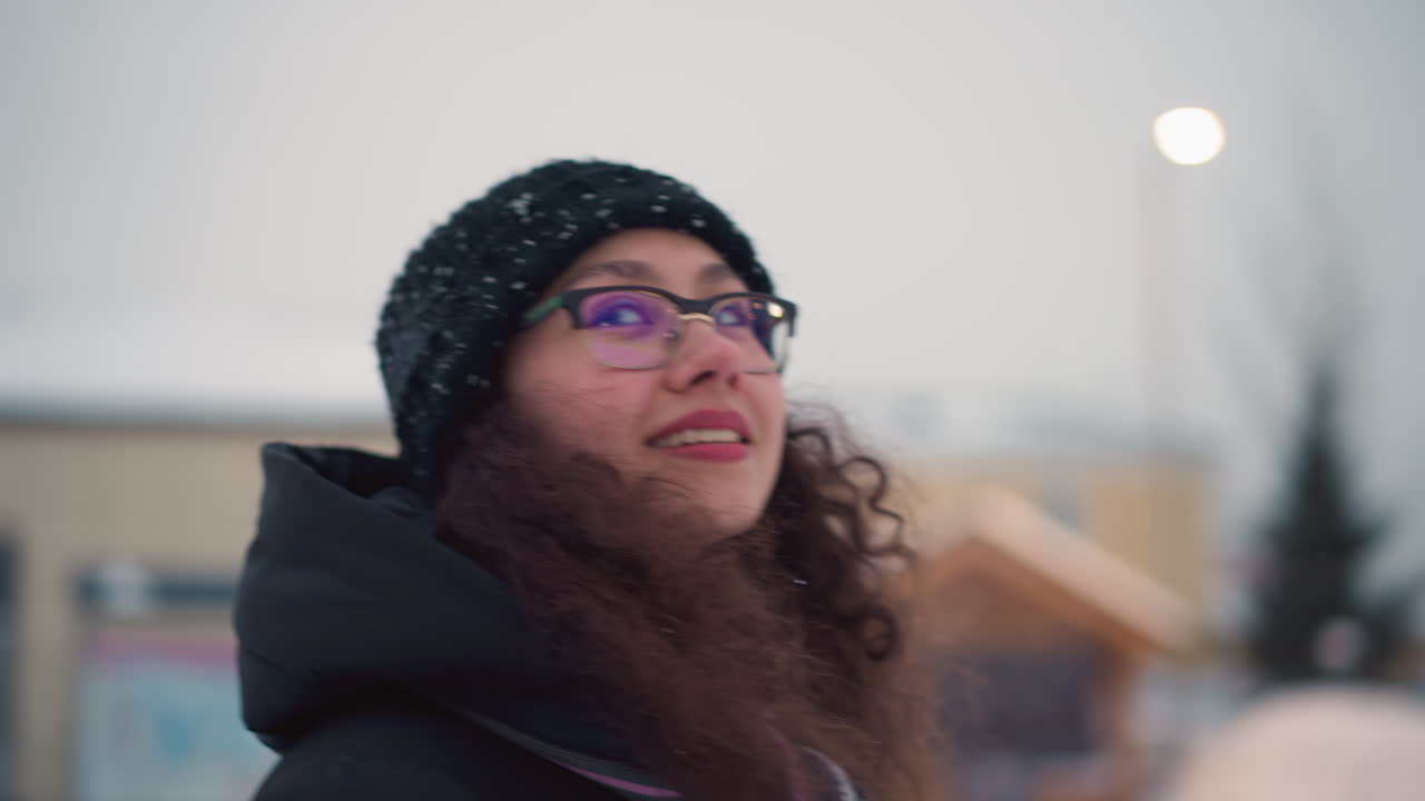 Uno skater con cappotto invernale nero e cappello di lana con fiocchi di neve è all'aperto in un parco divertimenti innevato, giostra colorata sfocata sullo sfondo, atmosfera invernale con aria gelida e fascino festivo della stagione fredda