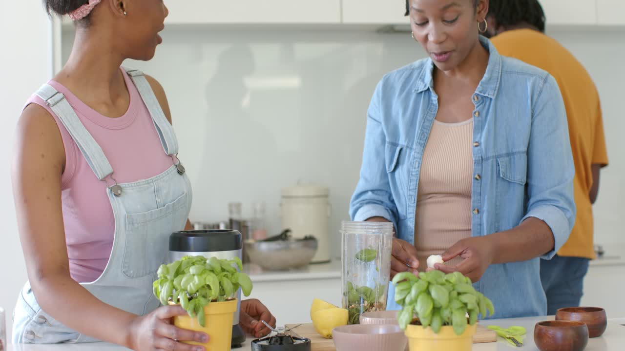 Female friends tearing basil and adding garlic into blender jar at kitchen island, preparing pesto