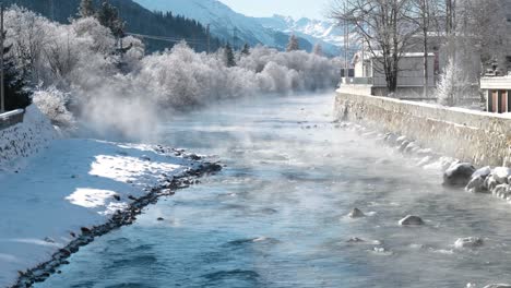 small stream of water flowing around frozen snow covered trees during winter with fog and mist