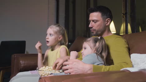 Vater-Und-Seine-Beiden-Kleinen-Töchter-Essen-Popcorn-Und-Schauen-Sich-Einen-Film-An,-Während-Sie-Nachts-Zu-Hause-Auf-Dem-Sofa-Sitzen-1