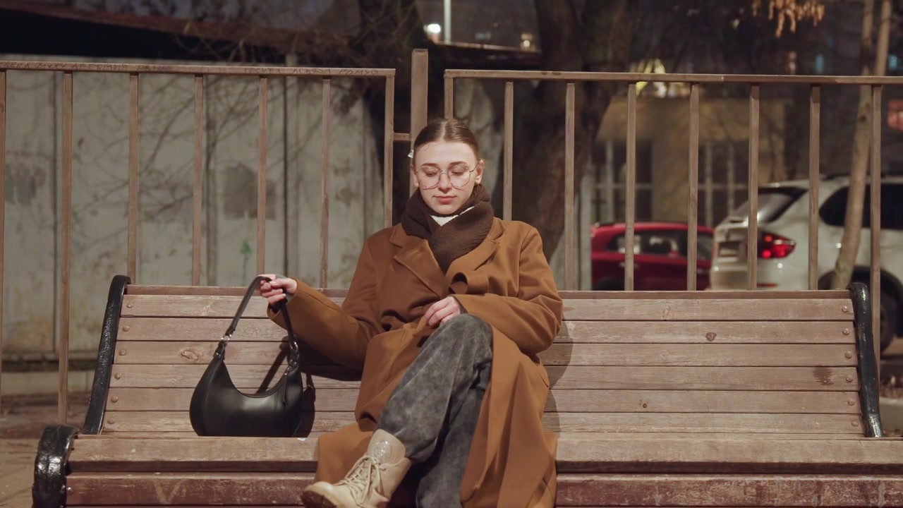 Donna caucasica seduta da sola su una panchina di sera, con cappotto marrone e occhiali, che posa la borsa accanto a sé, in postura contemplativa con uno sfondo di recinzione urbana e auto parcheggiate, una pausa calma e riflessiva durante il tragitto.