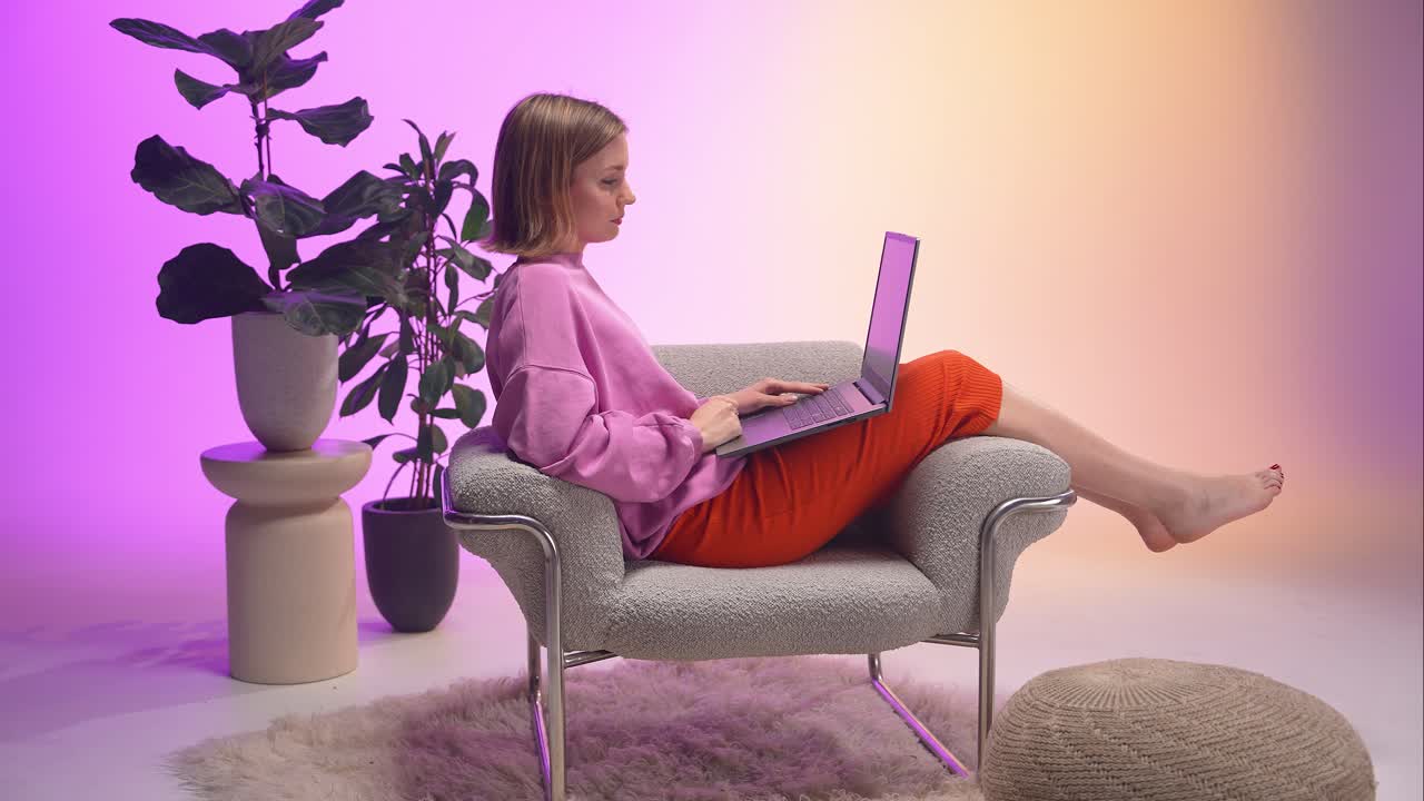 A woman in a pink sweatshirt and orange pants waves during a video call, sitting barefoot with a laptop. The cozy interior, soft rug, and gradient lighting reflect calm, digital connection and ease