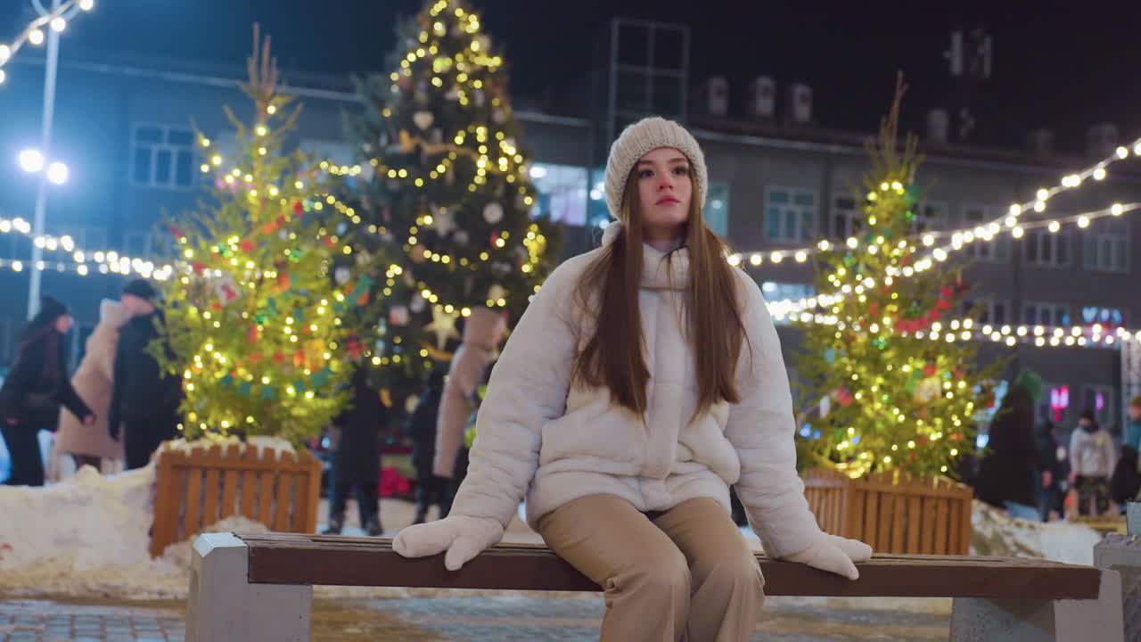 Woman in cozy winter outfit and beanie walks toward wooden bench in lively city at night, festive string lights illuminate background with people skating and socializing