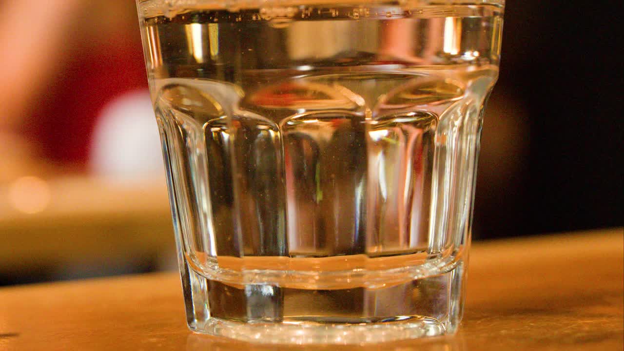 Hand pours clear water into glass, bubbles form, warm indoor lighting, close-up, steady camera