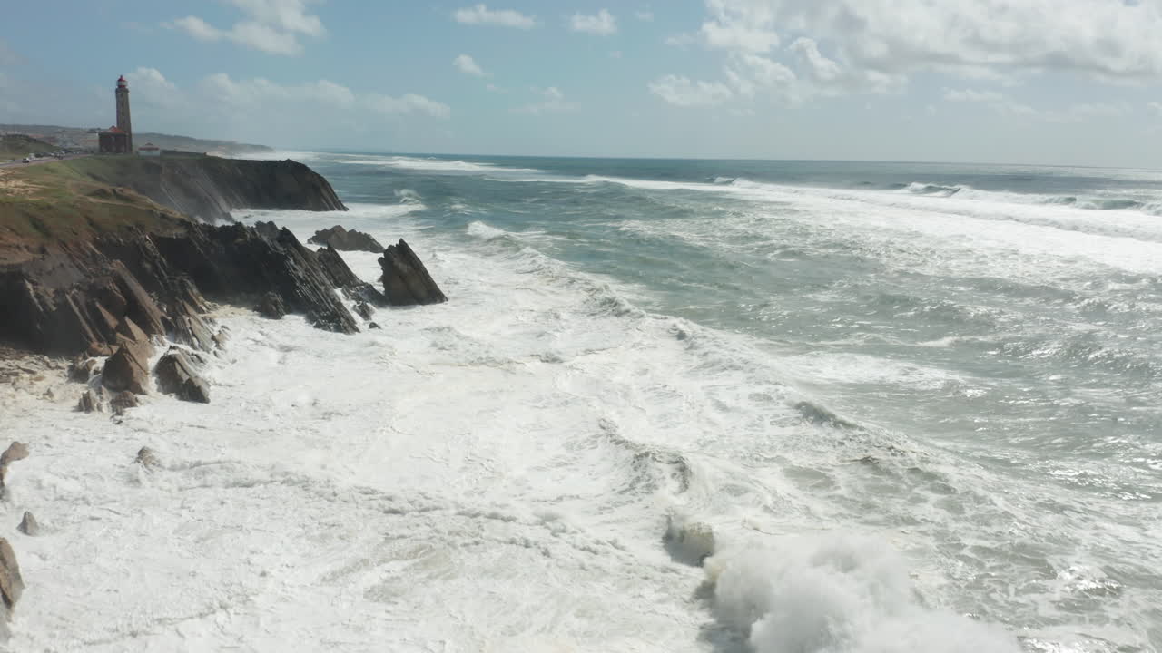 Flying over rough waves from sea slamming into rocky cliffs with a lighthouse in the background