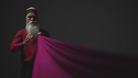 Fotografía-De-Estudio-Con-Iluminación-Tenue-De-Un-Hombre-Sikh-Mayor-Doblando-Tela-Para-Un-Turbante-Contra-Un-Fondo-Oscuro-Simple,-Filmada-En-Tiempo-Real