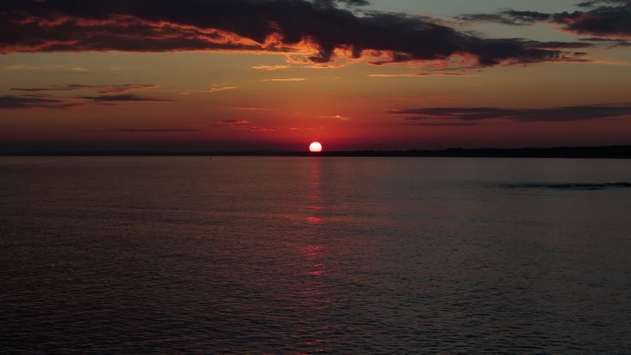 In Maldonado, Uruguay, a drone flies toward a glowing sunset over a calm sea. The sun reflects gently on the water, with colorful clouds painting the sky on a peaceful evening