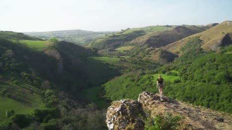 handheld, static wide shot of young blonde woman standing on the edge of a cliff above thor's cave, peak district, england at sunset