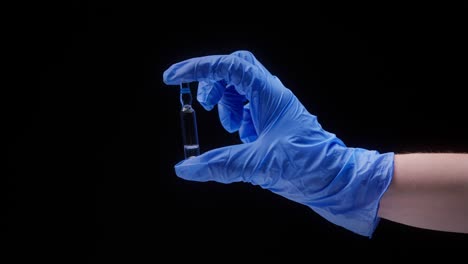 close-up of hand wearing blue medical glove, holding ampoule with vaccine against coronavirus. nurse showing sample of medicine on black background, vaccination against covid, laboratory research
