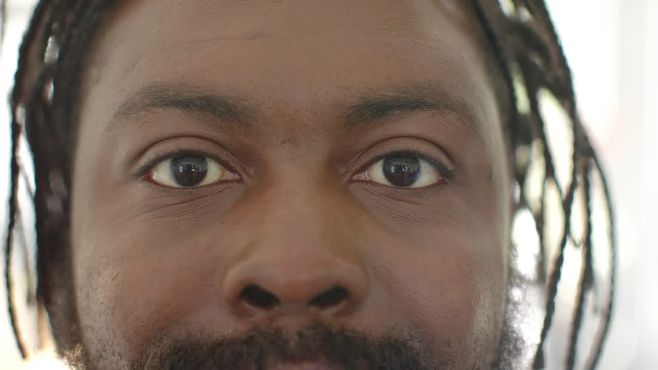 Uomo africano adulto, con dreadlocks, fissa lo sguardo in una stanza luminosa, gli occhi si addolciscono e forma un sorriso che mostra i denti.