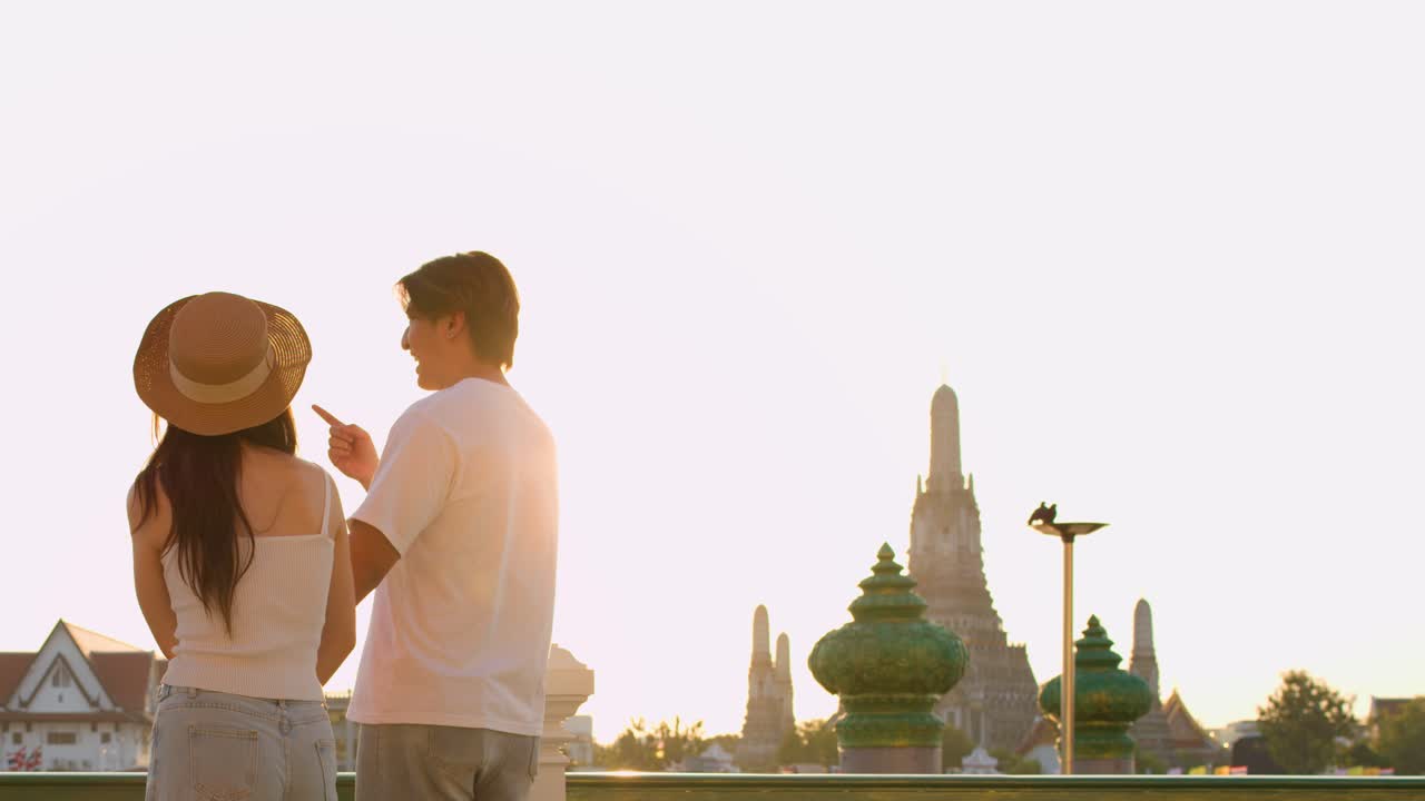 Young couple admires temple skyline at sunset, holding hands, soft backlight, wide shot