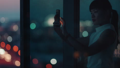 asian business woman using smartphone taking photo of city at night enjoying beautiful view working late in office