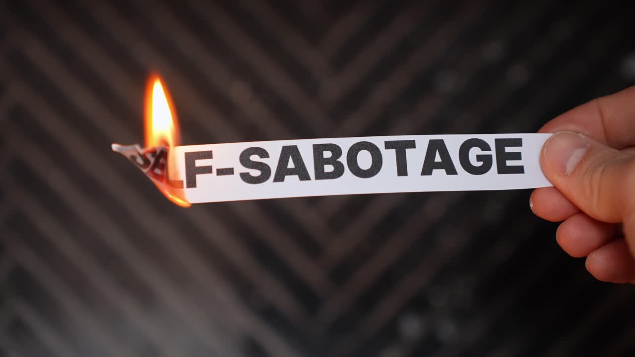 Mano brucia un'etichetta di carta con la scritta “AUTO-SABOTAGGIO”, a simboleggiare la rottura di schemi negativi – autodisciplina, fiducia in sé stessi e crescita personale. Sfondo scuro con spazio per il testo