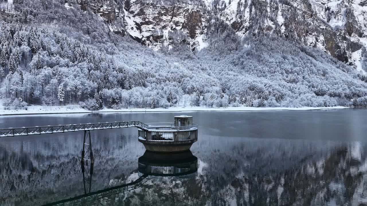 Winter scene of a circular reservoir tower connected by a narrow bridge on calm Klontal Lake surrounded by snow