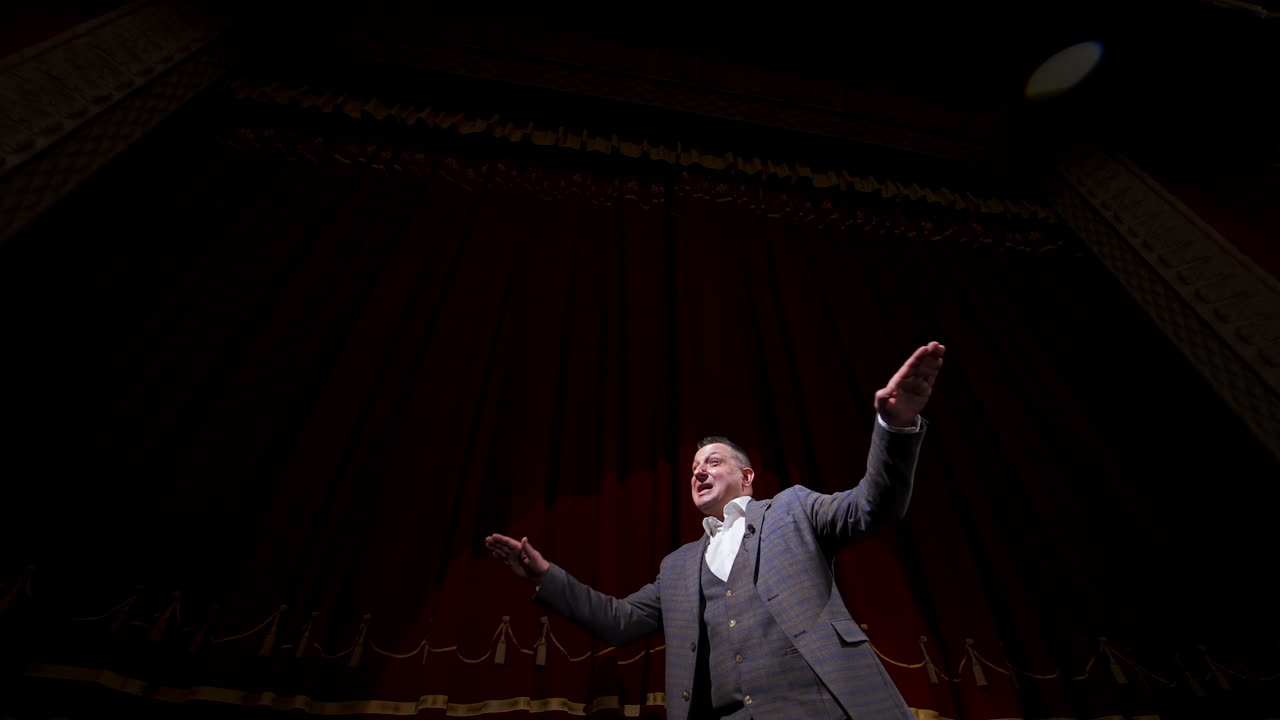 Theatrical man on stage. Performer in a suit moving on stage on red curtains background. Professional actor performing in theater. View from below.