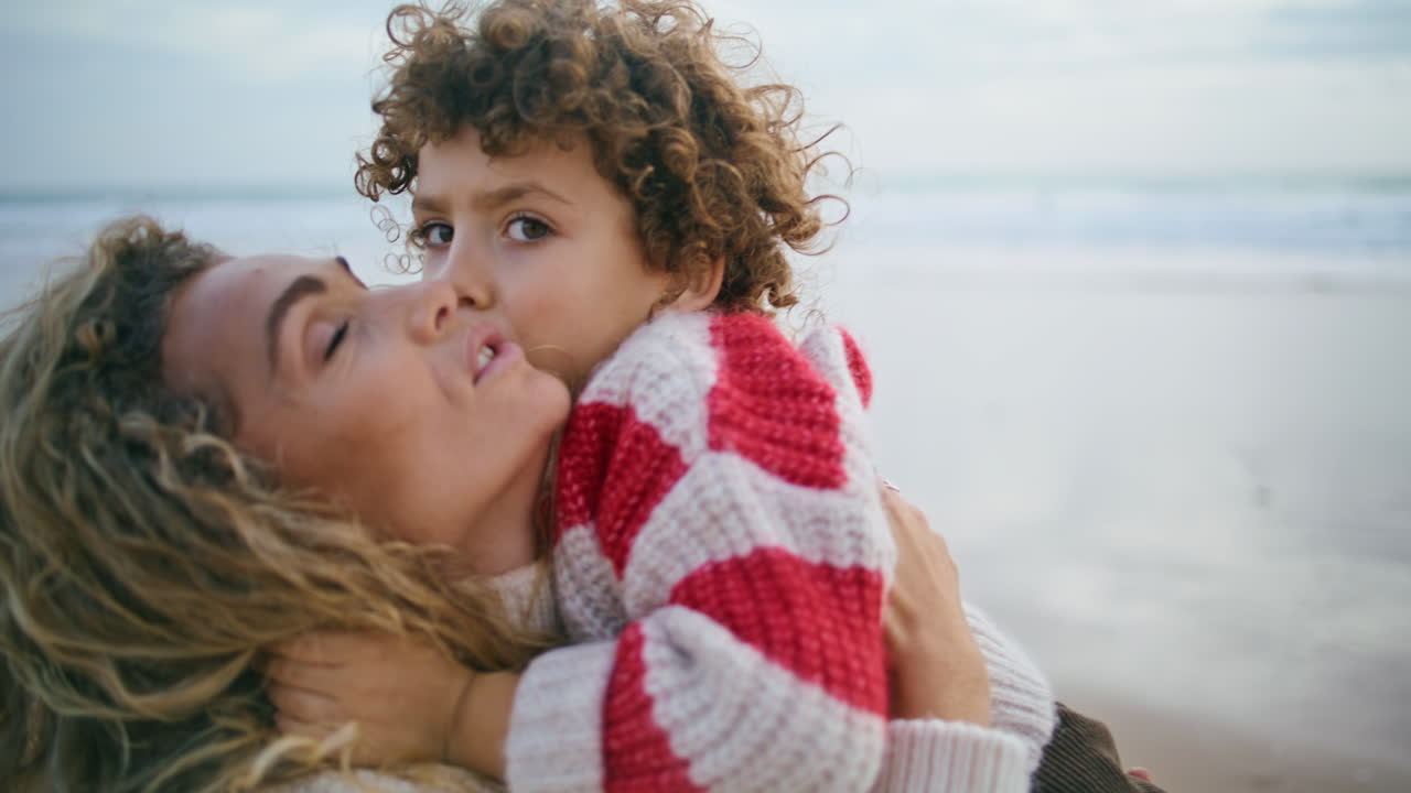 Madre felice che coccola il figlio sulla riva dell'oceano in autunno, primo piano. Emozioni familiari sincere.
