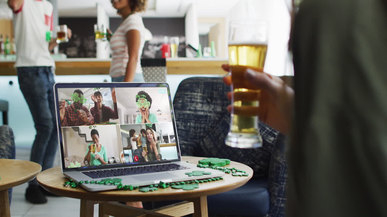 Gruppo eterogeneo di amici sorridenti con birra che indossano oggetti a forma di trifoglio in videochiamata sul laptop