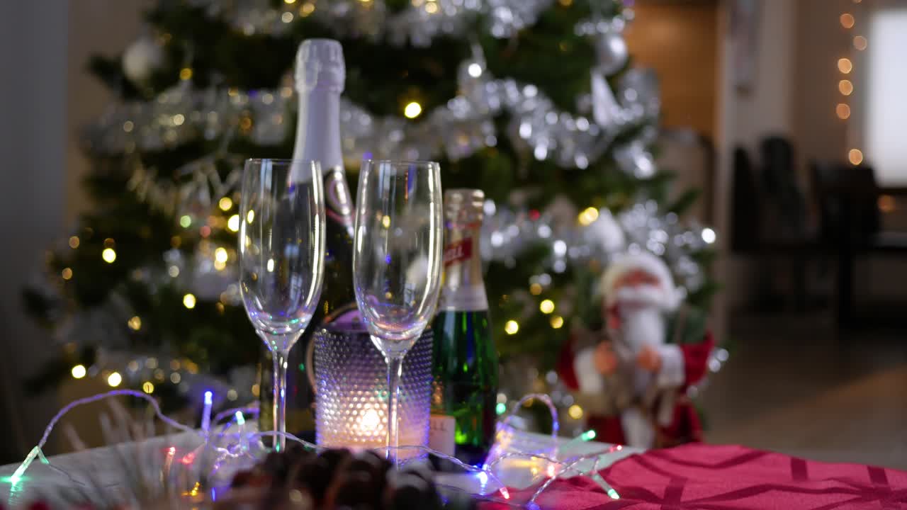 Due bicchieri vuoti e due bottiglie di champagne accanto a una candela accesa in un portacandele, sullo sfondo di un albero di Natale decorato.