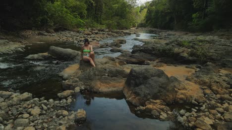 flyover model in bikini on rock in jungle river in costa rica, 4k drone