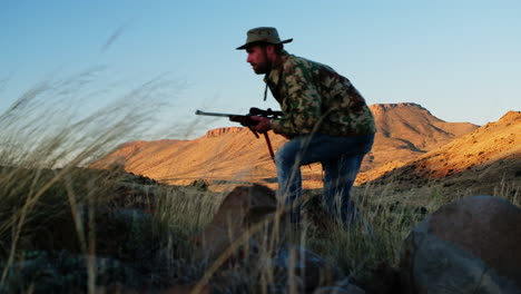 Hunter-with-rifle-walks-crouched-over-stalking-prey,-rocky-landscape,-profile