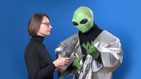 woman journalist in glasses with microphone interviews green skinned alien in silver clothes on blue chromakey background.