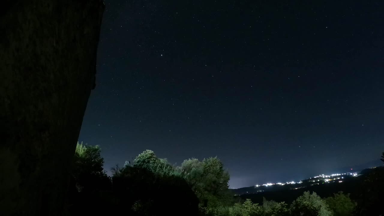 Time lapse di scie stellari nel cielo notturno sopra la silhouette della foresta e luci lontane della città con formazione rocciosa