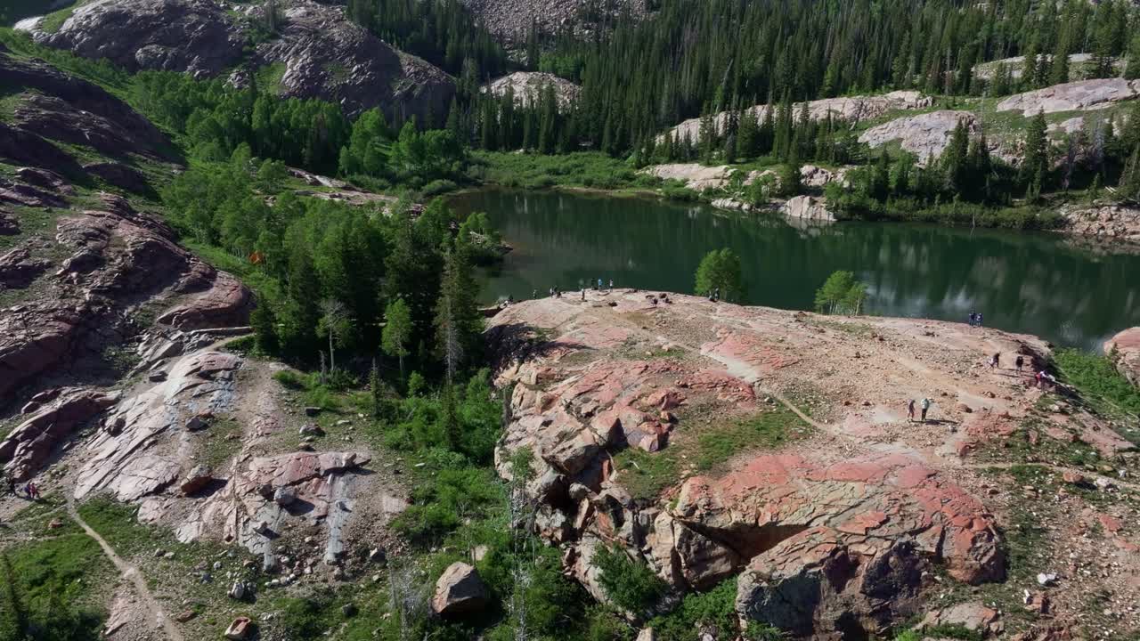 Aerial drone tilting up shot revealing Lake Blanche in Big Cottonwood Canyon near Salt Lake City, Utah, on a bright green summer morning with rocky peaks, lush foliage, and Sundial Peak towering above