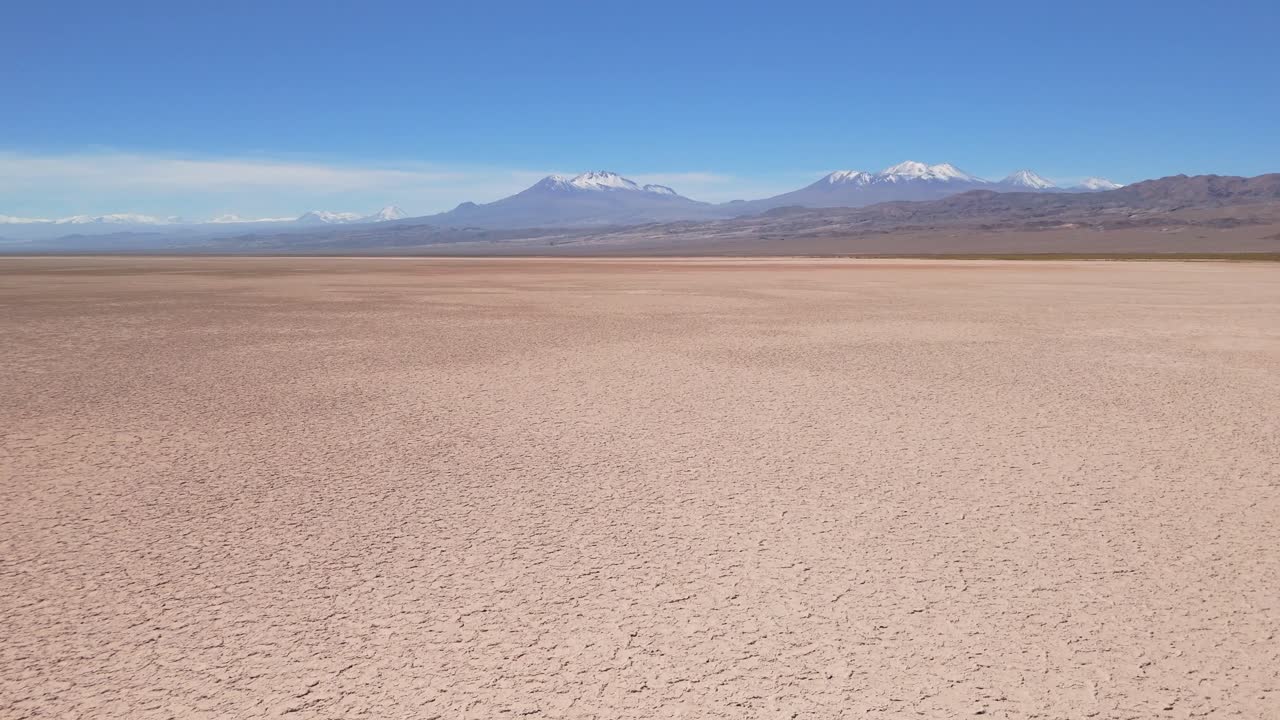 High-altitude drone shot over the Atacama Desert salt flat, showing salt, earth, and lithium crusts under daylight, with snow-capped Andes mountains in the background.