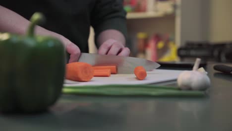 Close-up-footage-of-a-person-chopping-carrots-on-a-cutting-board-in-a-kitchen,-with-a-green-bell-pepper-and-garlic-visible-in-the-foreground