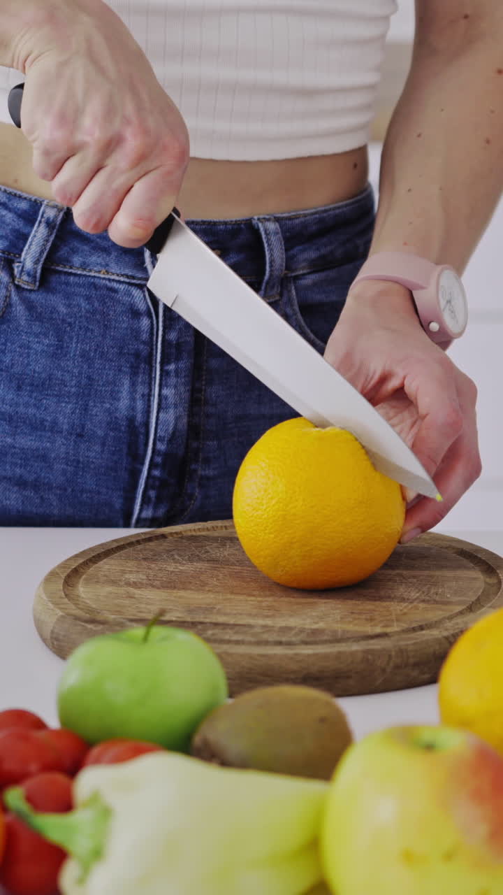 Woman with sharp knife cutting orange into two halves. Table full of fresh fruit and vegetables in the kitchen. Woman preparing healthy food for losing weight. Vertical video