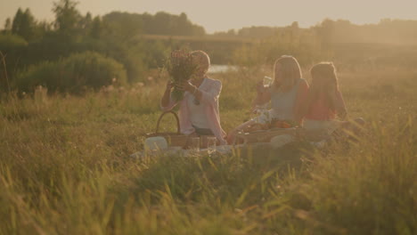 elder sister picking flowers from basket while mother and daughter sit on picnic mat drinking water on sunny day, family enjoying outdoor picnic surrounded by lush greenery