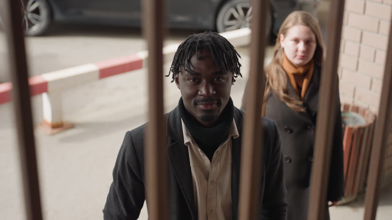 En los fotogramas de la cámara se ve a dos amigos a través de una rejilla de hierro en la entrada de un edificio; el hombre mira a la cámara con expresión serena, mientras que la mujer permanece detrás, ligeramente desenfocada; la escena mezcla líneas, texturas y luz tenue.