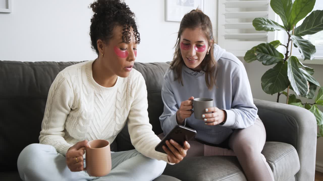 Em close-up, amigas diversas usando patches para os olhos, navegando no celular e tomando café em casa
