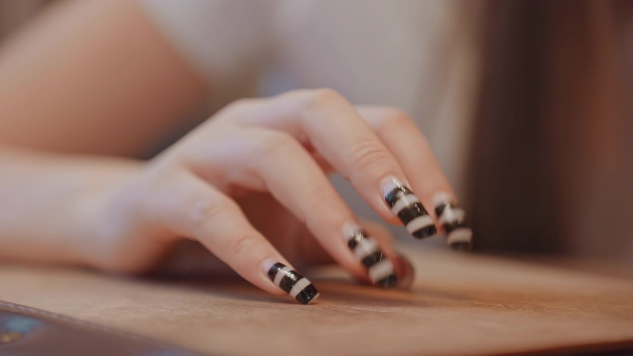 Mano in primo piano con unghie nere a strisce che riposa su un tavolo di legno, influencer di bellezza in studio, illuminazione cinematografica calda, smalto lucido, movimento delicato delle dita, dettaglio di design delle unghie raffinato.