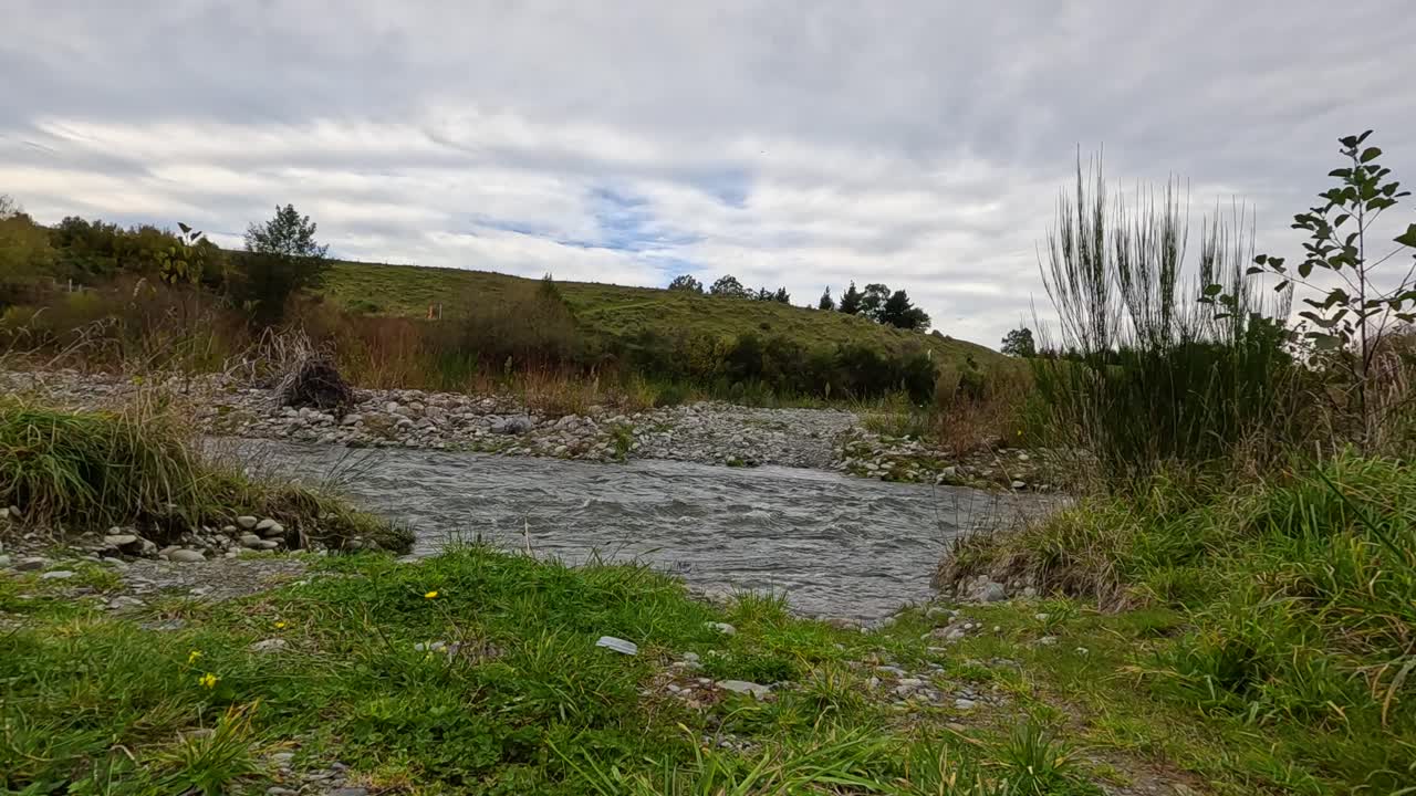 Camera steadily advances across grassy terrain toward a shallow, rocky stream under overcast skies, capturing natural textures and tranquil autumn landscape