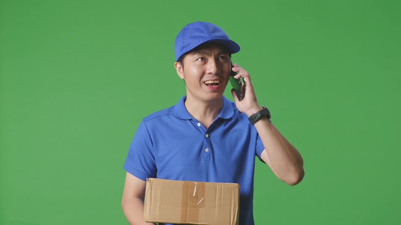 Close Up Of Asian Male Courier In Blue Uniform Walking And Talking On Smartphone While Delivering A Carton On Green Screen Background In The Studio