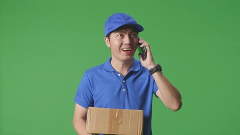 primer plano de un mensajero masculino asiático en uniforme azul caminando y hablando en un teléfono inteligente mientras entrega un cartón en el fondo de la pantalla verde en el estudio