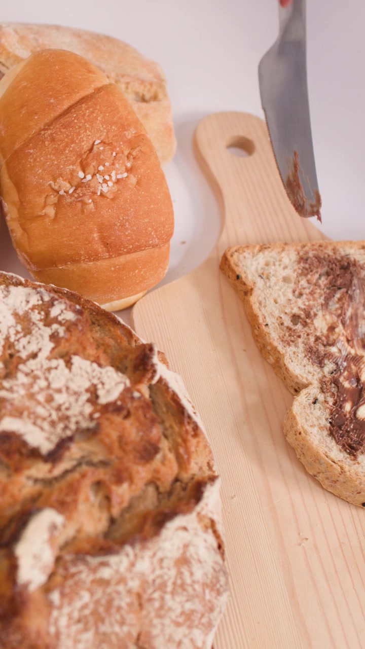 A hand spreads creamy nut butter onto multigrain bread beside sourdough and crusty baguettes.